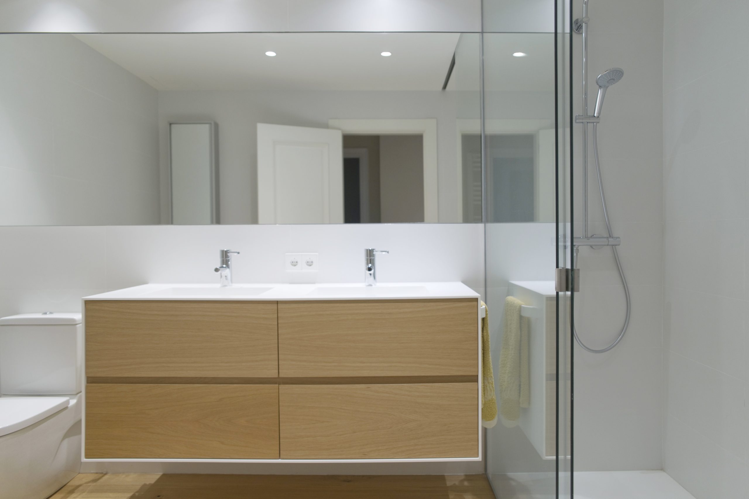 Proyecto de Reforma íntegra de baño en una vivienda de Pamplona (Navarra). Paredes y techo en blanco y suelo en madera de haya. Lavabo doble y mampara de vidrio. Un espacio cálido y limpio.