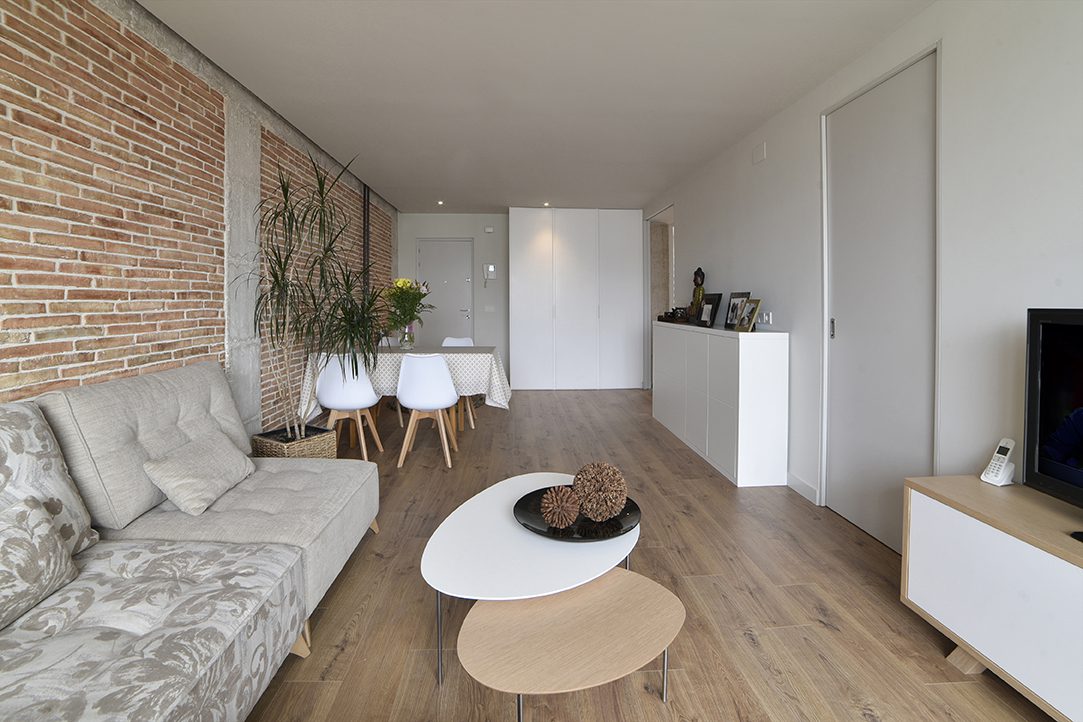 Proyecto de reforma integral de una vivienda completa en el barrio de San Juan de Pamplona (Navarra). Vista de la sala de estar decorada con ladrillo rojo a la vista y pilares y vigas de hormigón que combina con el suelo cerámico imitación madera y las paredes pintadas en blanco.