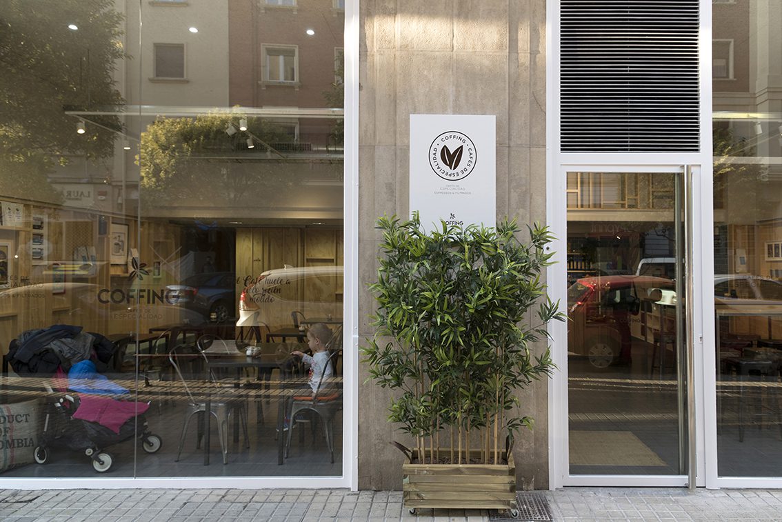 Proyecto de reforma integral de local comercial para la Cafetería Coffing en Pamplona (Navarra). Decoración industrial que combina sillas y mesas metálicas de color oscuro con paredes de madera clara que emulan el tabique de madera. Espacio luminoso por sus dos ventanales y amplio.