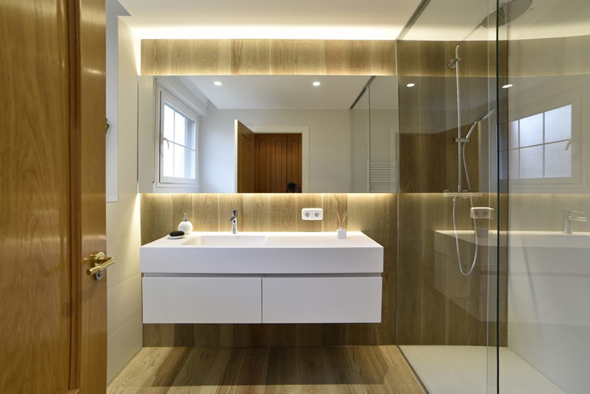 Proyecto de Reforma íntegra de baño en Gorráiz (Navarra). Luz indirecta, lavabo suspendido en color blanco, pared y suelo en cerámica imitación madera y paredes y techo en cerámica blanca. Diseño lineal, limpio y actual.