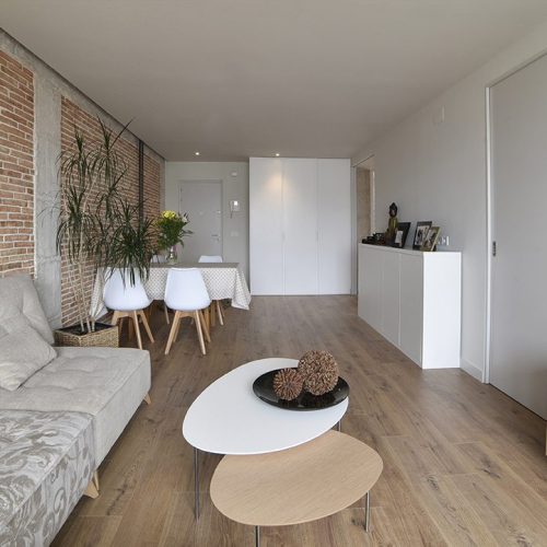 Proyecto de reforma integral de una vivienda completa en el barrio de San Juan de Pamplona (Navarra). Vista de la sala de estar decorada con ladrillo rojo a la vista y pilares y vigas de hormigón que combina con el suelo cerámico imitación madera y las paredes pintadas en blanco.