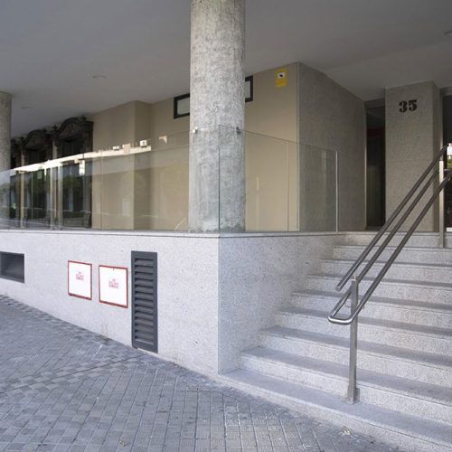 Proyecto de rehabilitación de portal y ascensores de un edificio de viviendas en Pamplona (Navarra). Exterior del portal reformado con pilares al aire, escaleras con pasamanos y barandilla de vidrio.