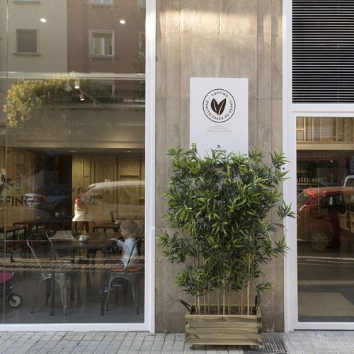 Proyecto de reforma integral de local comercial para la Cafetería Coffing en Pamplona (Navarra). Decoración industrial que combina sillas y mesas metálicas de color oscuro con paredes de madera clara que emulan el tabique de madera. Espacio luminoso por sus dos ventanales y amplio.