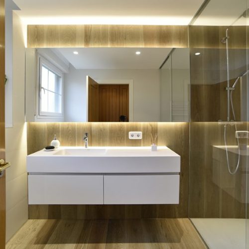 Proyecto de Reforma íntegra de baño en Gorráiz (Navarra). Luz indirecta, lavabo suspendido en color blanco, pared y suelo en cerámica imitación madera y paredes y techo en cerámica blanca. Diseño lineal, limpio y actual.